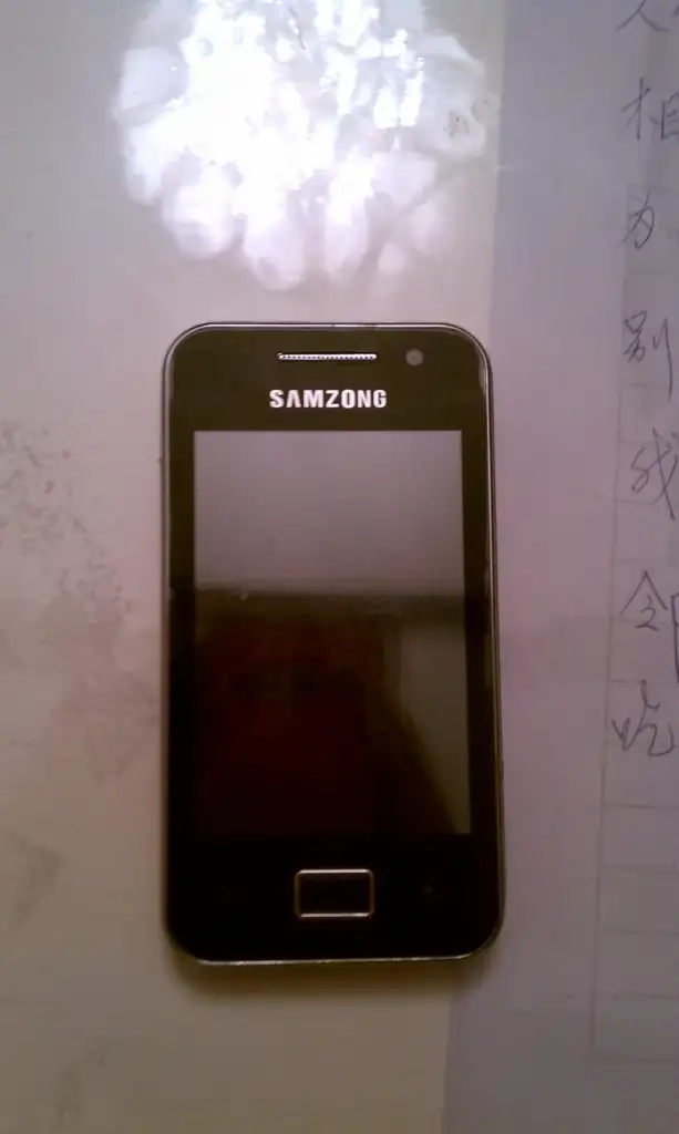 A smartphone with "SAMZONG" branding is placed on a surface. Its screen is off, and writing in a different language is visible on a paper to the right.