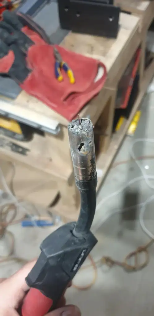 Close-up of a frayed and damaged electrical cable with exposed wires. In the background, a workbench with red cloth, pliers, and various tools is visible. The setting appears to be a workshop.