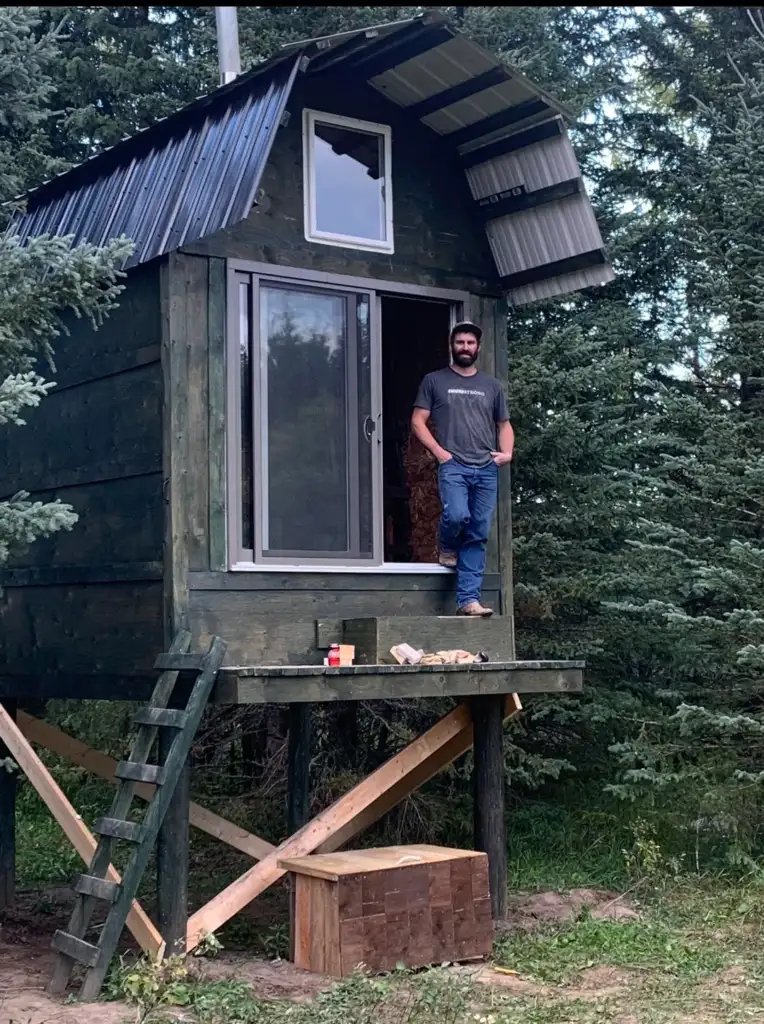 A person stands on the deck of a small, elevated cabin surrounded by evergreen trees. The cabin has a metal roof, a large sliding door, and a ladder for access. There are tools and wooden planks nearby.