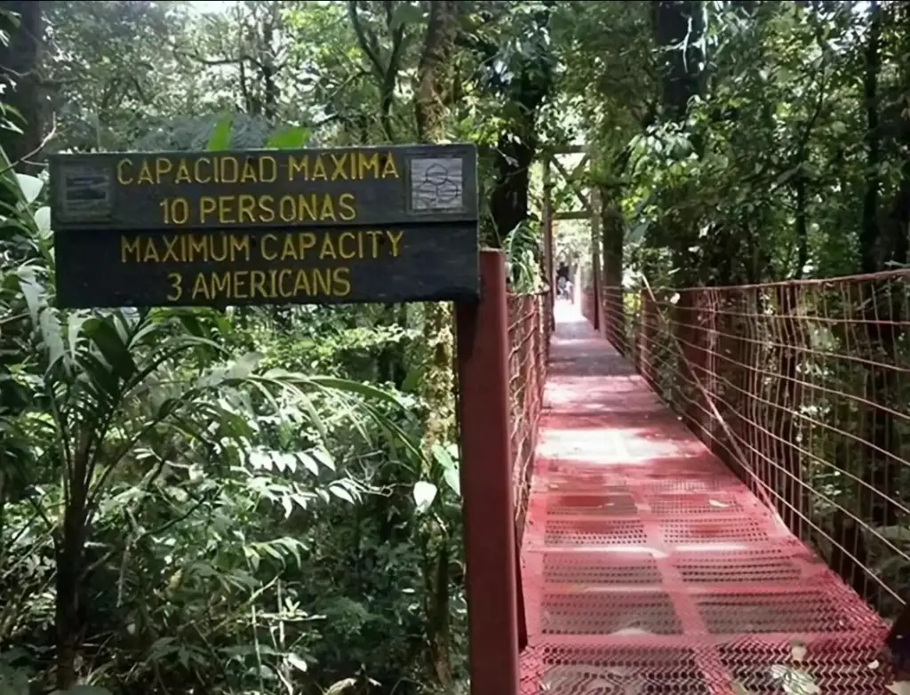 A red metal bridge in a lush forest. A nearby sign reads "Capacidad Maxima: 10 Personas, Maximum Capacity: 3 Americans." The bridge is surrounded by dense greenery on both sides.