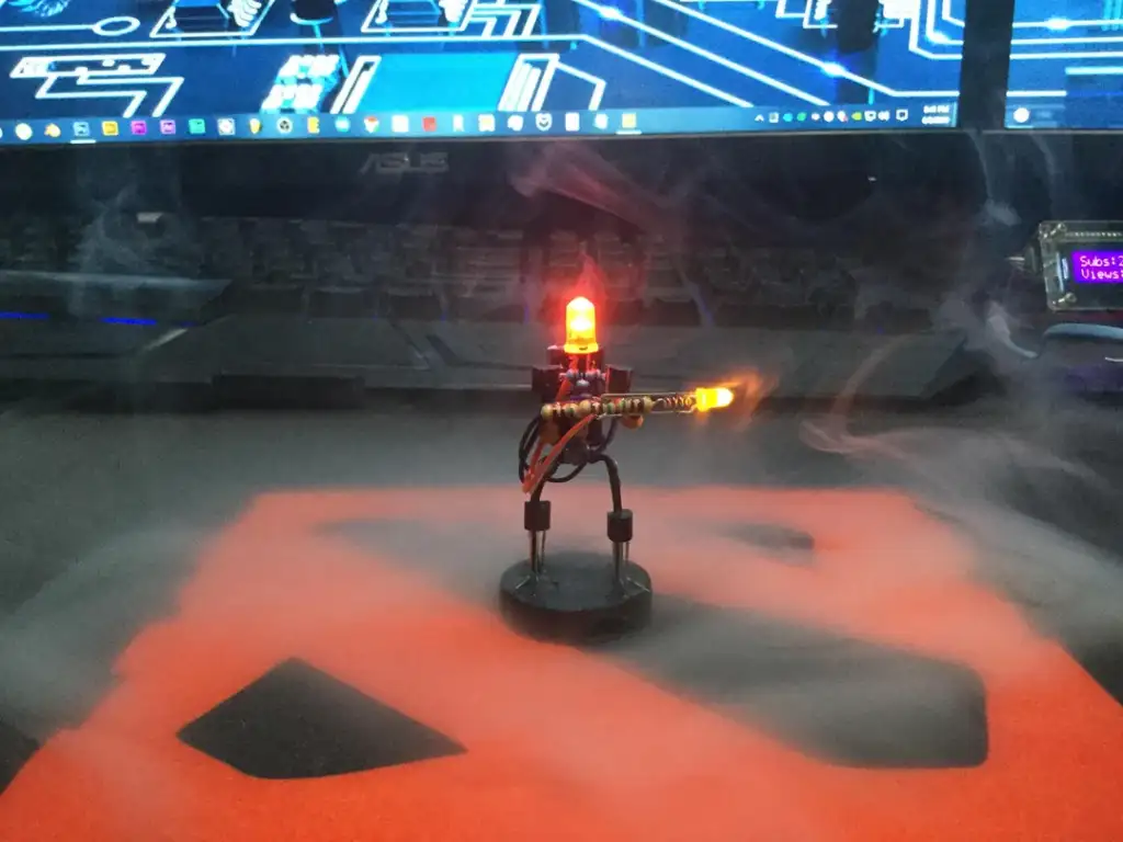 A small robot figure with a glowing red LED head and a yellow LED on its arm stands on a table. Smoke surrounds the robot, with a computer monitor displaying a blue circuit board design in the background.