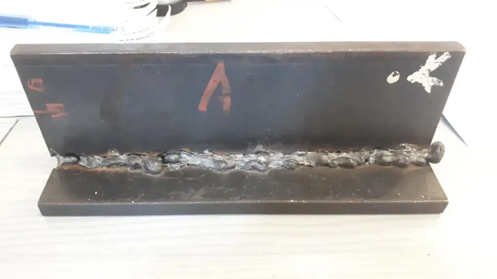 A metal piece displaying a welded seam runs horizontally across the center. The surface has visible weld marks and irregularities. The piece is positioned upright on a surface, with letters and numbers marked in paint on the side.