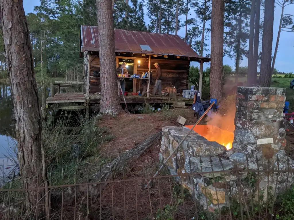 A rustic wooden cabin with a tin roof stands near a serene lake, surrounded by tall pine trees. Two people are on the porch with camping gear. In the foreground, a stone fire pit with a bright fire burns, adding warmth to the outdoor setting.