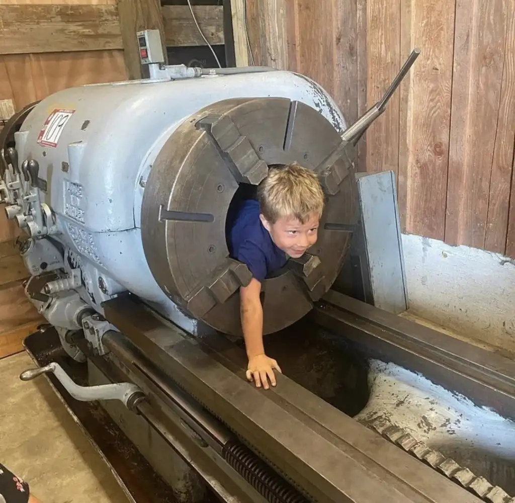 A child playfully crawls through the large opening of an industrial lathe in a workshop setting. The child is emerging headfirst with a curious expression. The machinery and wooden walls provide a rustic backdrop.