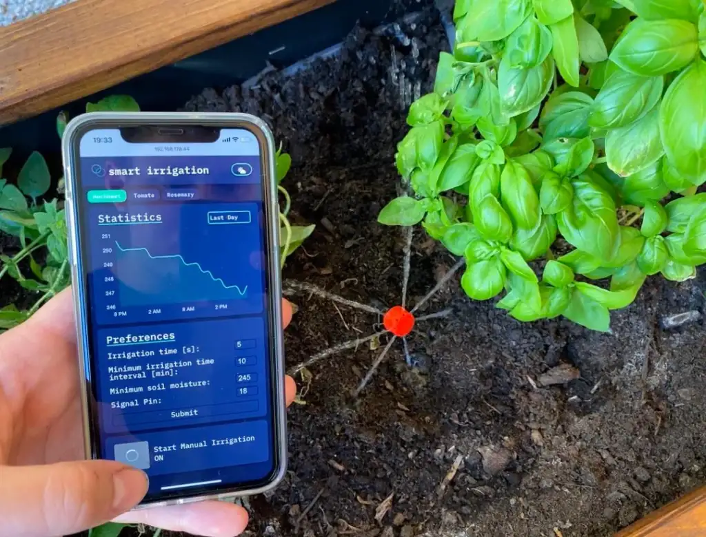 A person holds a smartphone displaying a smart irrigation app next to a basil plant. The app shows statistics and preferences for watering, while a red irrigation sensor is visible in the soil.