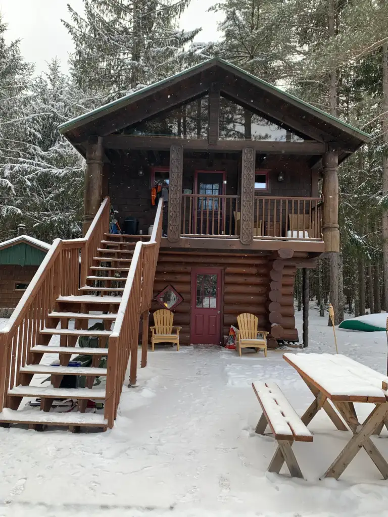 A log cabin with a front porch and an upper balcony, surrounded by snow-covered trees. Wooden stairs lead to the balcony, and there are two Adirondack chairs on the porch. A wooden picnic table is in the foreground on the snow.