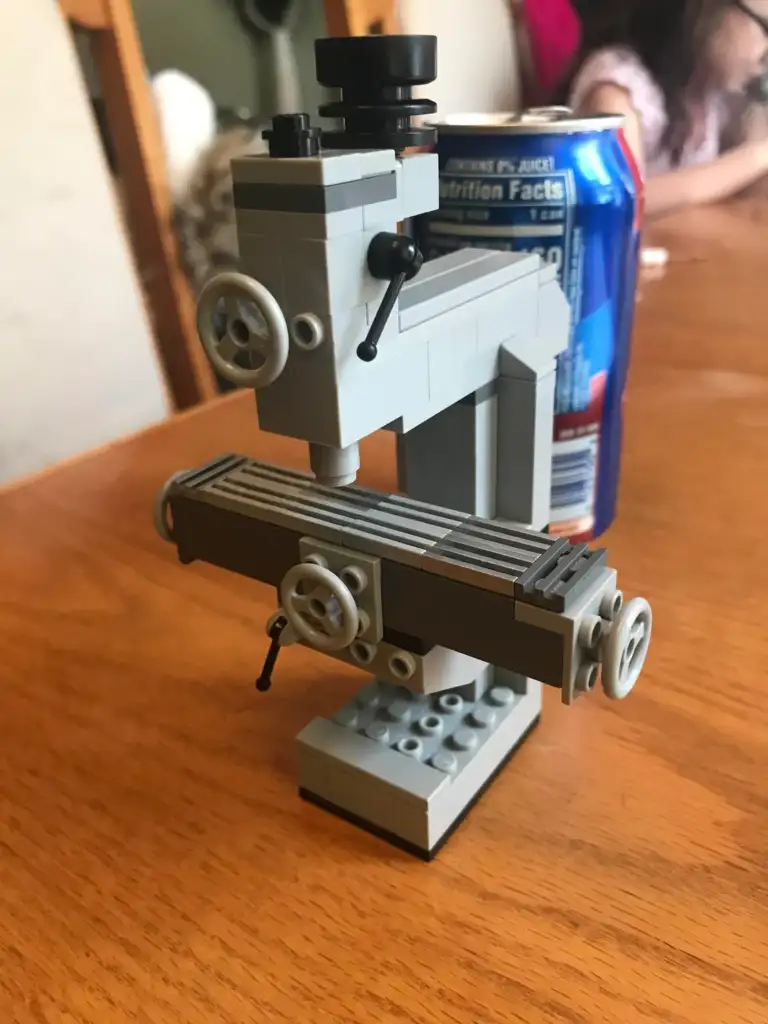 A miniature milling machine model made from LEGO bricks is displayed on a wooden table. Behind it is a can of soda, and a person is partially visible in the background.