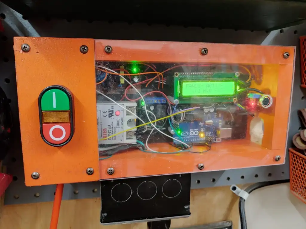 An orange control box with a transparent cover displays electronic components and wires. It features a green button for power on and a red button for power off. A small screen shows dynamic text and green LED lights are illuminated inside.