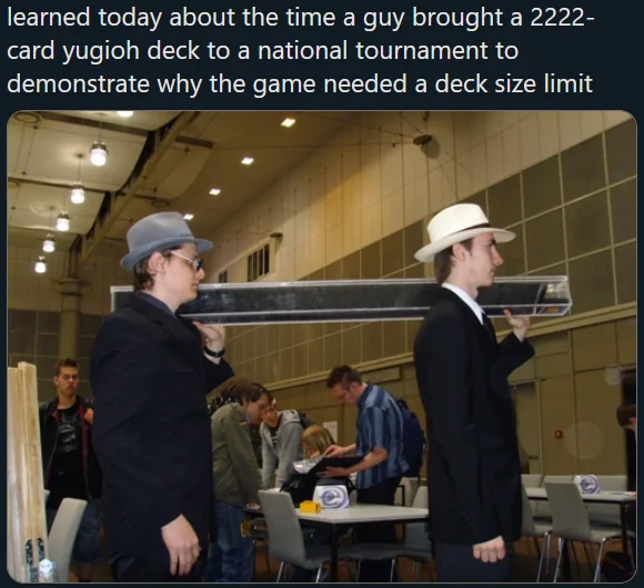 Two people in suits and hats carry a large, long box, presumably containing a 2222-card Yu-Gi-Oh deck, through a tournament venue. Curious onlookers watch as they walk through a well-lit room with tables and chairs.