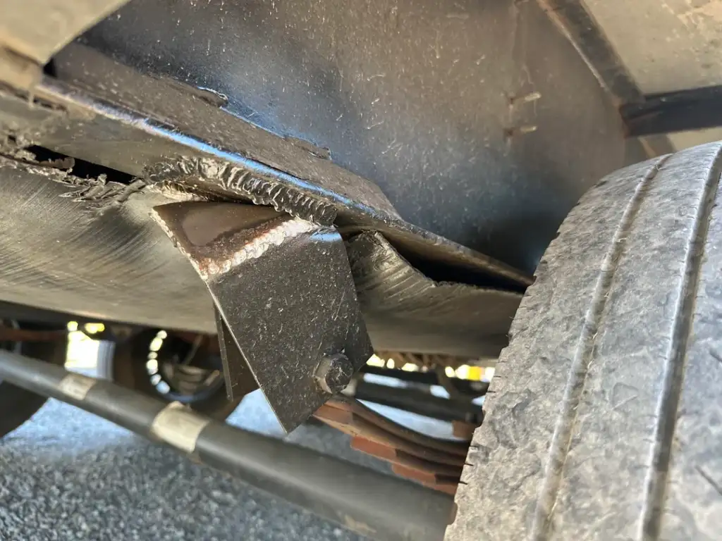 Close-up of a vehicle's undercarriage showing a cracked metal bracket and a worn tire. The image highlights visible damage to the metal, with surrounding mechanical parts in the background.