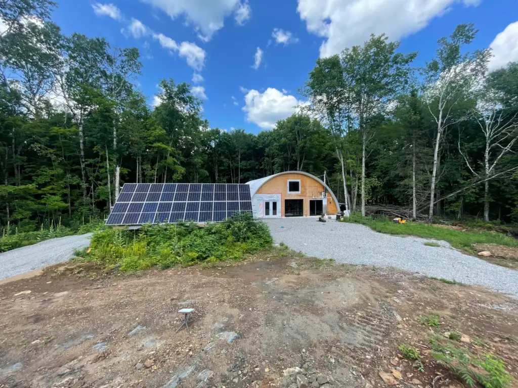 A modern, semicircular wooden house with large windows and a gravel driveway is nestled in a forested area. Solar panels are installed on the ground nearby. The sky is blue with scattered clouds.