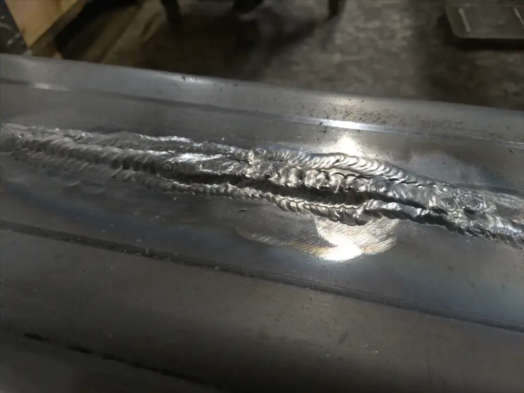 Close-up of a metal surface featuring a freshly welded seam with visible ripples and texture. The weld is situated along the center of the surface, reflecting light with subtle iridescence. The background shows a dim workshop setting.