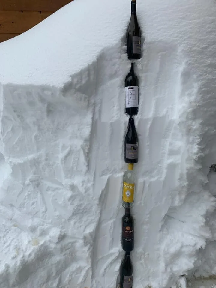 Four wine bottles are embedded vertically in a deep snowbank, with the labels facing forward. The snow is packed tightly around the bottles, holding them in place.