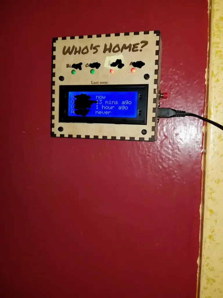 A wooden home status board mounted on a red wall displays the question "Who's Home?" with a blue digital screen showing the last seen times of different individuals, indicated by buttons labeled A, B, C, and D.