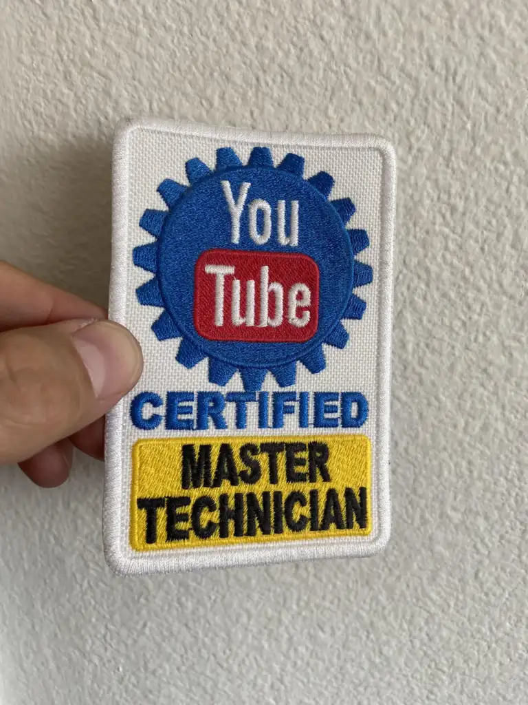 A hand holds a patch featuring a gear icon with the YouTube logo inside. Below, it reads "CERTIFIED MASTER TECHNICIAN" in blue and yellow text on a white background.