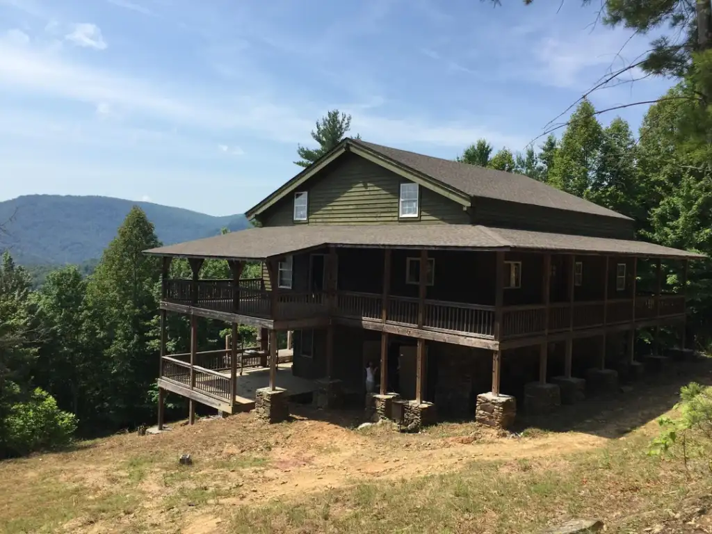 A large, rustic mountain cabin surrounded by trees with a wraparound porch. The house is set against a backdrop of tree-covered hills under a clear blue sky.