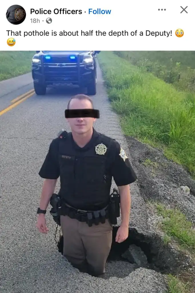 A police officer stands in a large pothole on a rural road, with a police vehicle parked behind. The pothole is deep enough to reach the officer's waist, illustrating its size.