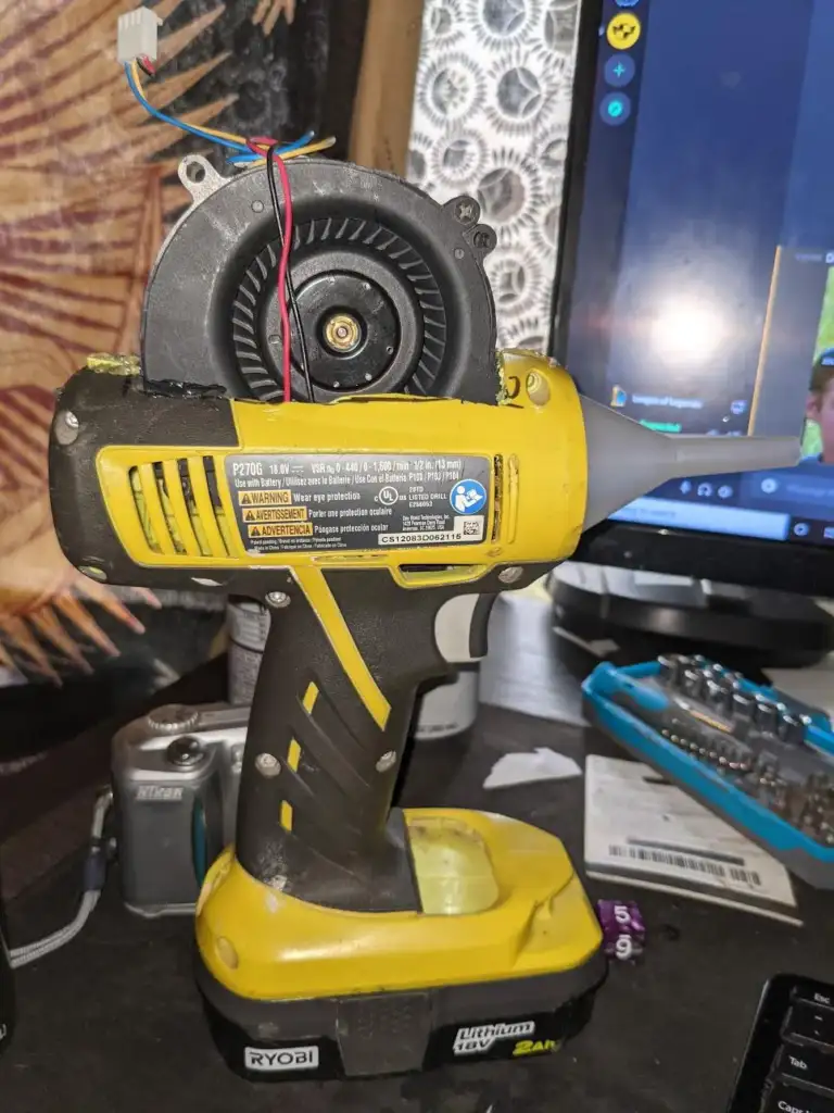 A yellow battery-powered tool modified with an attached small fan on top. The setup is placed on a cluttered desk with various items, including a camera, a monitor, and a small tool set in the background.