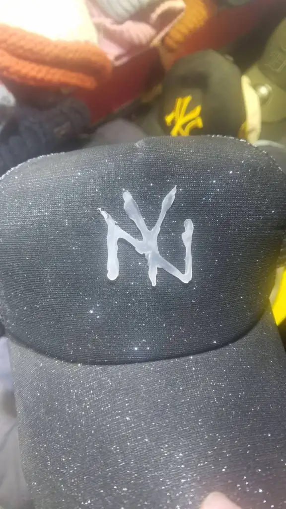 A black cap with a glittery texture has white glue dried on the front, forming an abstract design resembling "NY" lettering. Other caps are visible in the background.