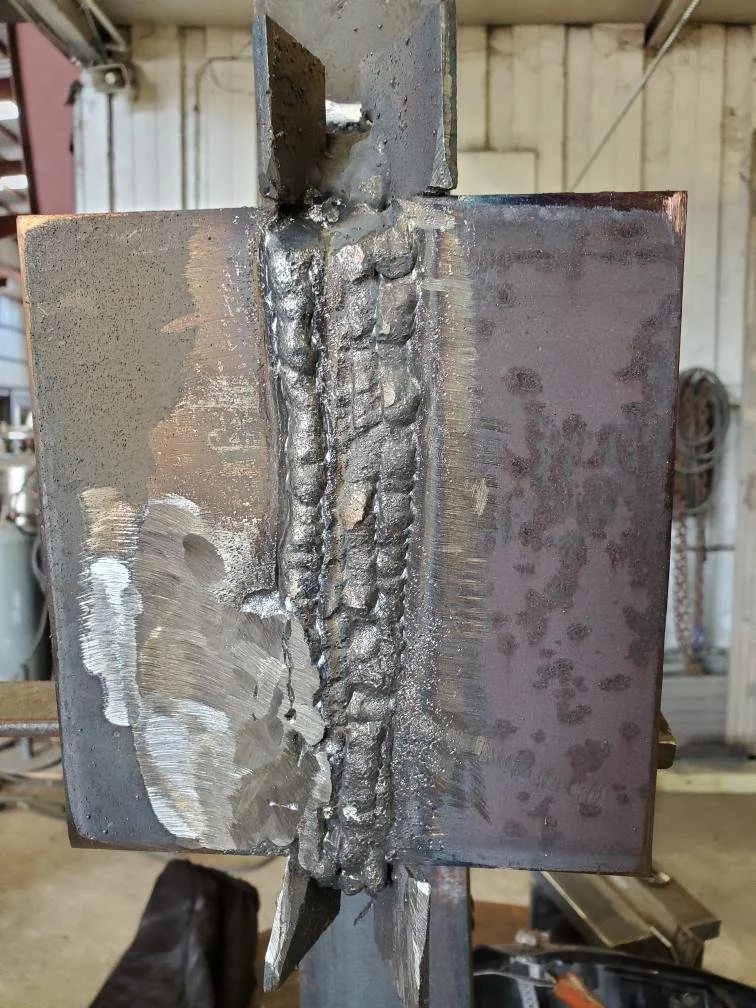 Close-up of a metal surface showing a rough weld seam. The weld is surrounded by a partially smoothed area on the left, with visible grinding marks. The background features an industrial setting with blurred tools and equipment.