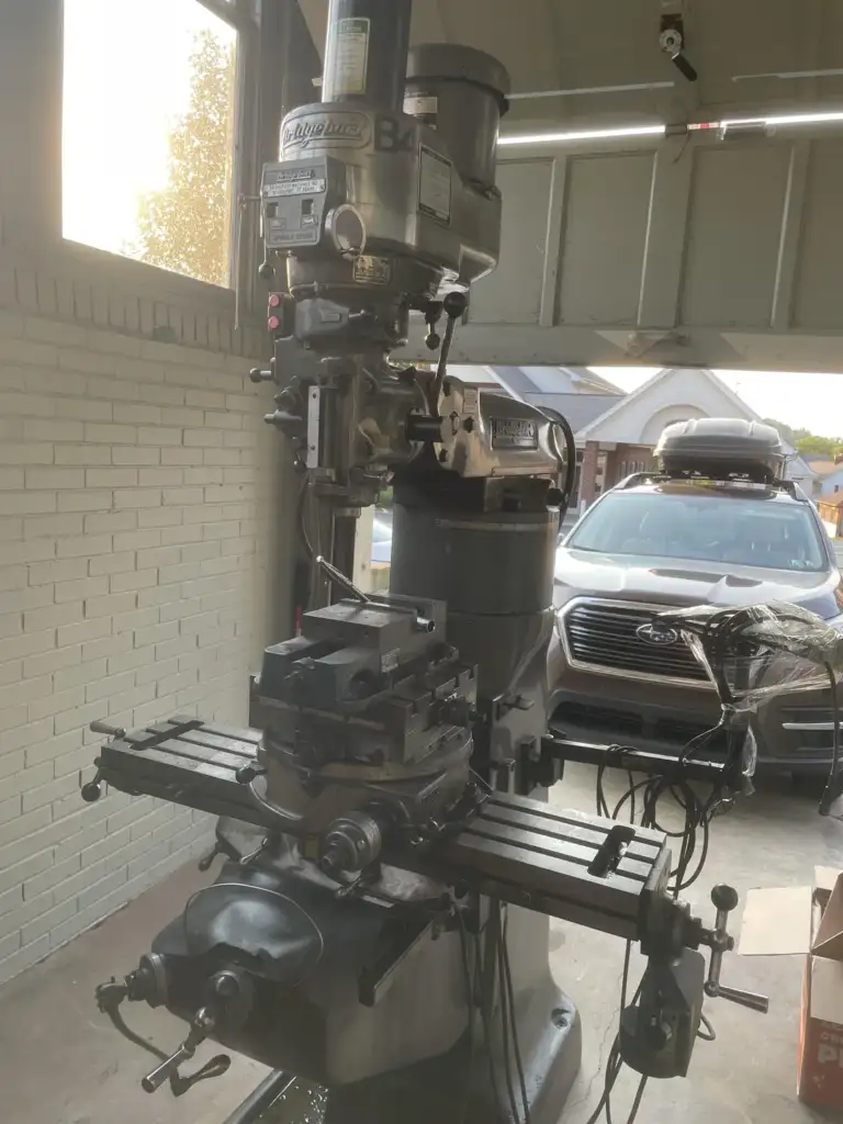 A milling machine is positioned inside a garage, with sunlight streaming through a window. A car is parked outside the open garage door, and boxes are visible nearby. The setting is a residential area.