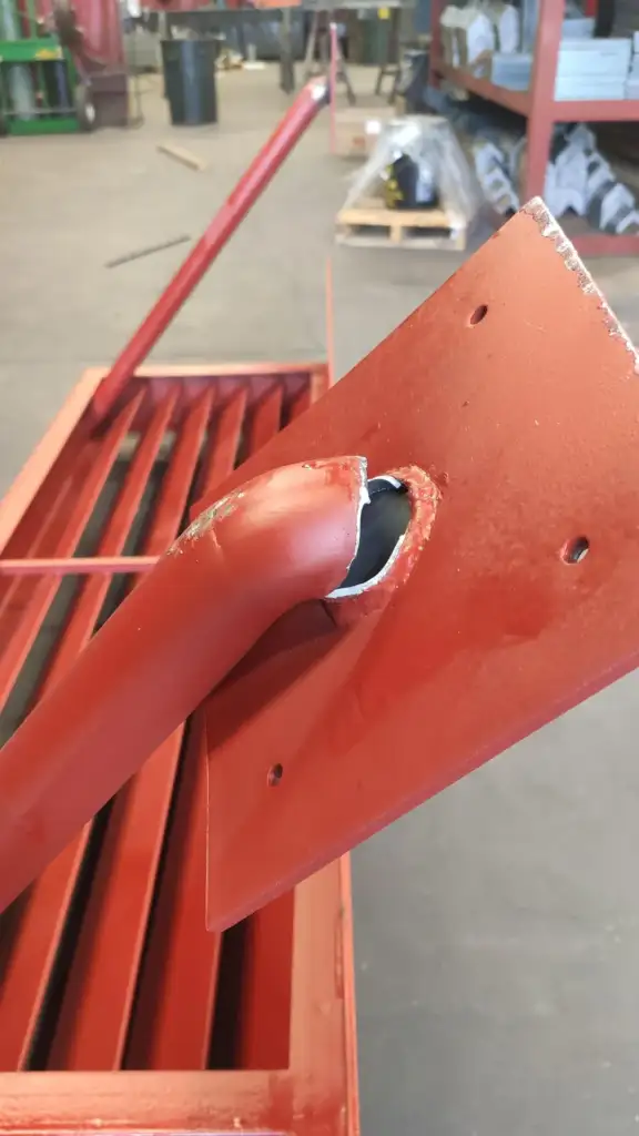 A close-up of a red metal structure, where a square plate is welded to a curved pipe. The joint shows rough welding marks and is slightly off-center. The structure is surrounded by shelves and materials in what appears to be a workshop.