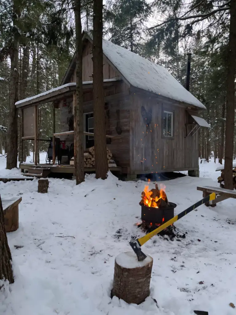 A rustic wooden cabin in a snowy forest, with a crackling fire burning in a metal fire pit nearby. Logs and a wooden bench are placed around the pit, creating a cozy outdoor seating area. Smoke rises gently into the wintry sky.