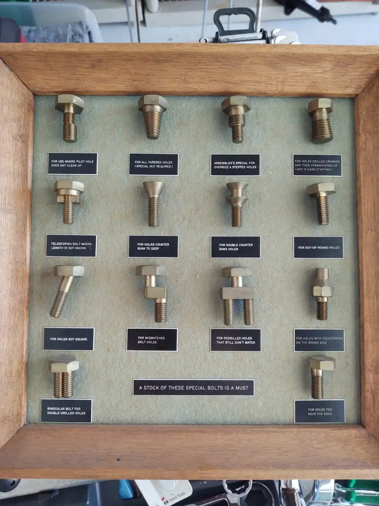 A wooden-framed display showcasing different types of bolts mounted on a board. Each bolt is labeled, highlighting its specific purpose, such as for broken nails, worn holes, countersink head, and more.