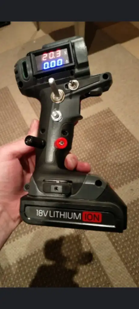 A hand holding a custom-made power drill with a digital display showing "20.3V" and "0.00". The drill is 18V lithium ion powered and has wires and modifications visible. It is held over a patterned carpet.