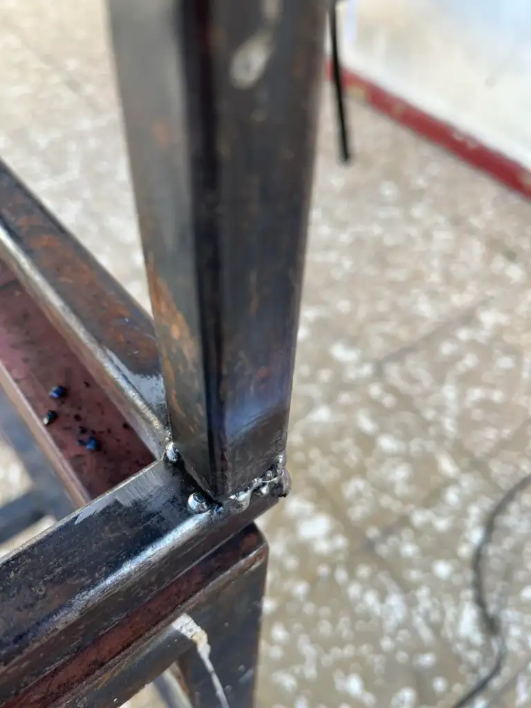 Close-up of a welding joint on a metal frame with visible weld beads. The metal appears aged with some rust. The background features a speckled, tiled floor.