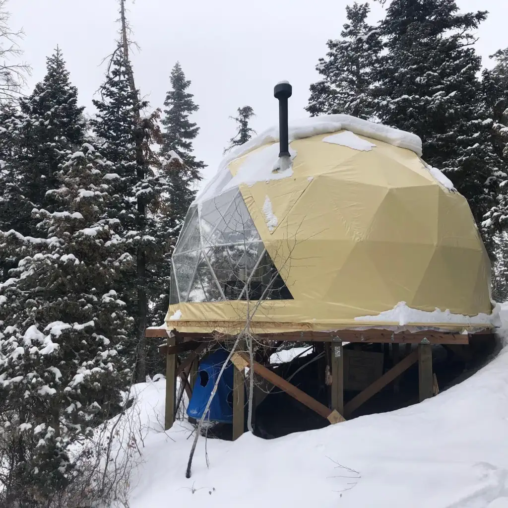 A geodesic dome covered in snow on a wooden platform in a forested area. The dome is partially transparent and surrounded by tall, snow-laden evergreen trees under a cloudy sky.