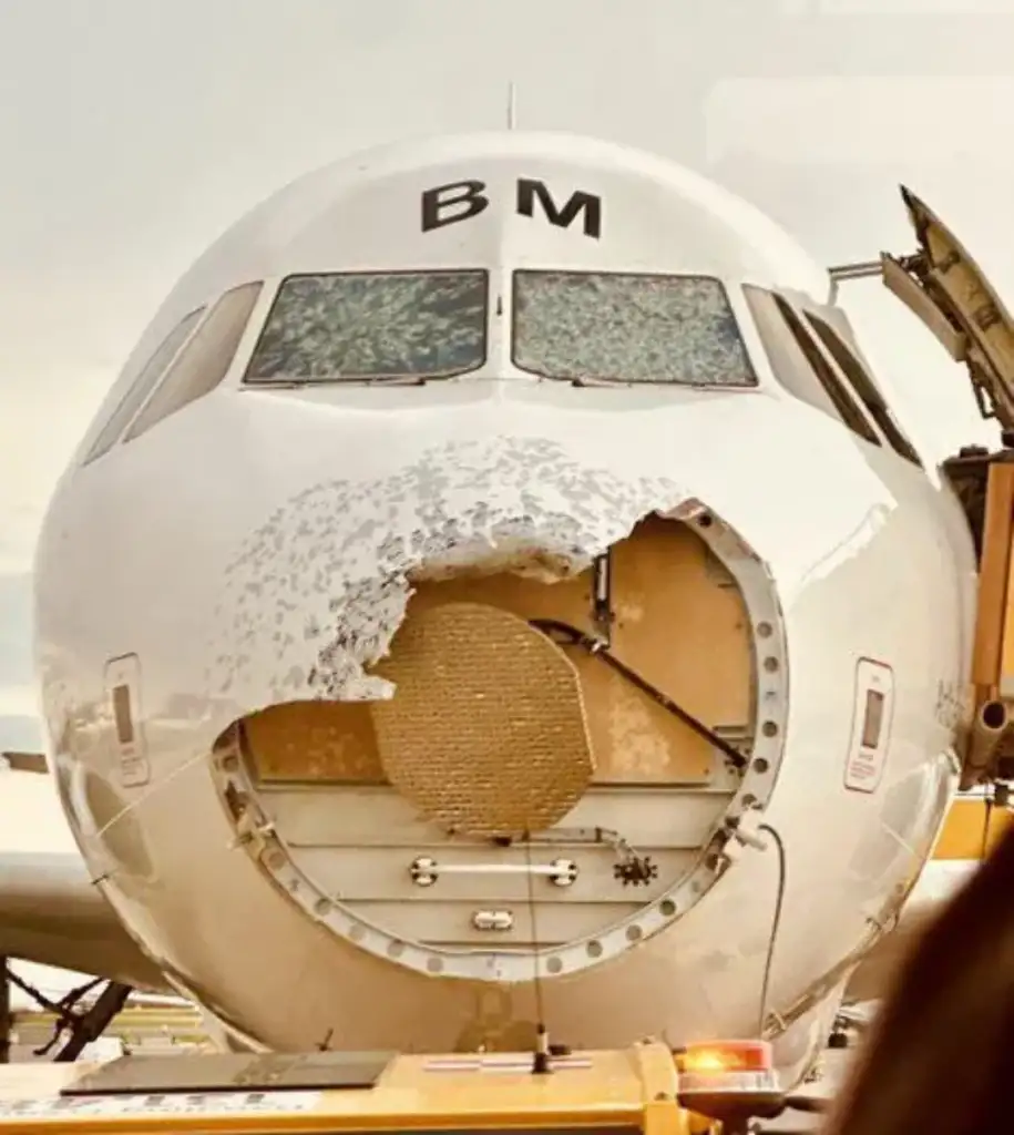 The front of an airplane with significant damage and a missing nose cone. The exposed radar and internal components are visible, and the cockpit windows appear intact. The aircraft is stationary, possibly at an airport.