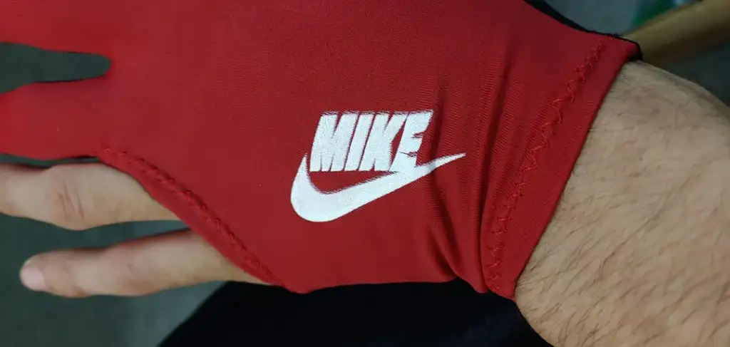 A right hand wears a red glove with the letters "MIKE" and a Nike swoosh logo. The glove covers most of the hand except the fingertips. The background is blurred and neutral in color.