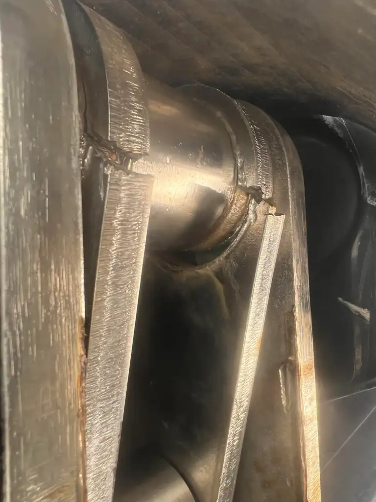 Close-up of a shiny, metallic pipe joint with visible welding marks. The metal surfaces have a textured appearance, reflecting light in certain areas. The joint is slightly corroded, showing signs of wear.