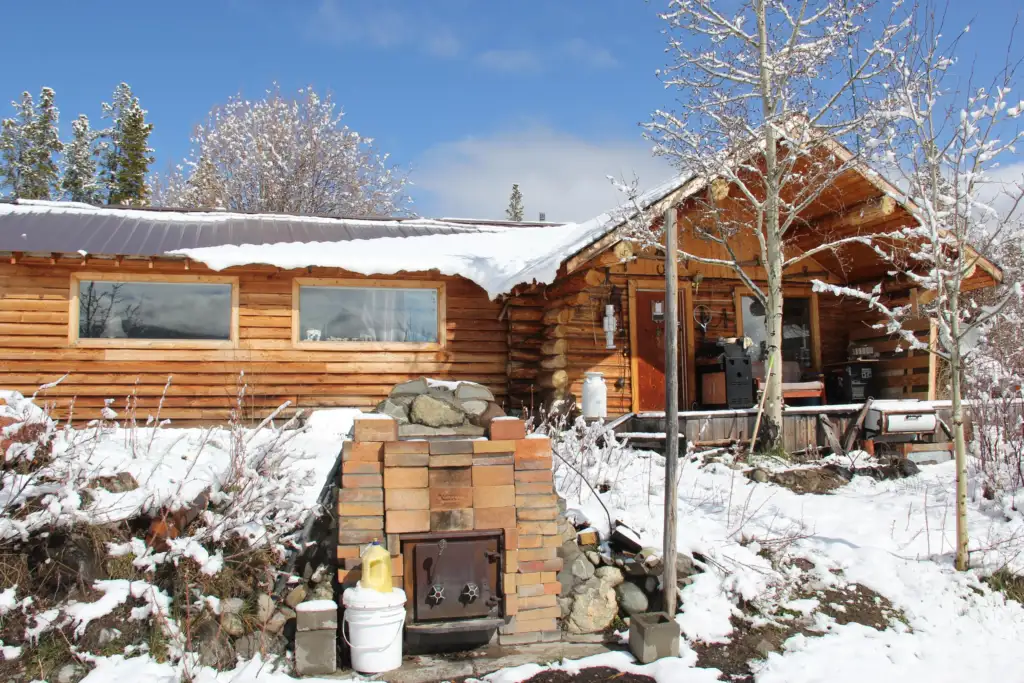 A rustic log cabin with a snow-covered roof is nestled in a snowy landscape. Bare trees and a brick outdoor oven are visible in the front yard. The blue sky is partially covered with clouds in the background.