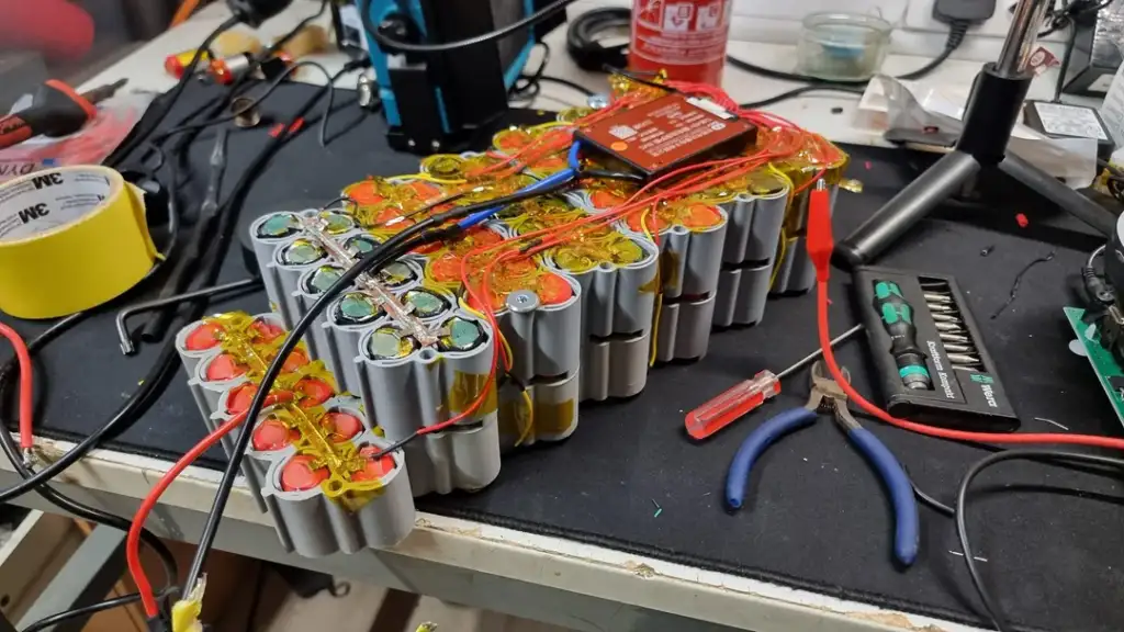 A cluster of connected cylindrical batteries with red and yellow wires arranged on a cluttered workbench. Tools, wires, and equipment surround the battery pack, along with a roll of yellow tape and a pair of pliers.