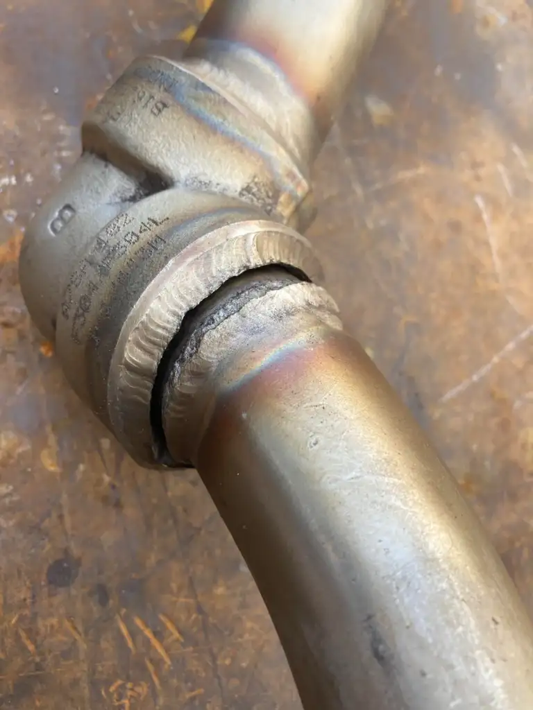 A close-up of a metal pipe with a large crack at the welded joint, showing discoloration from heat treatment. The background is a rusty, textured surface, suggesting industrial or workshop surroundings.