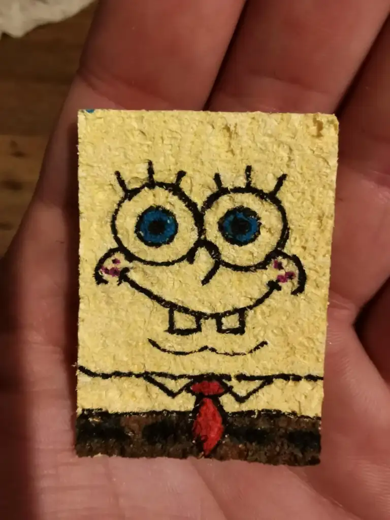 A hand holding a small, textured tile with a drawing of a smiling cartoon character in a white shirt and red tie. The tile is painted yellow with the character's face outlined in black with large blue eyes.