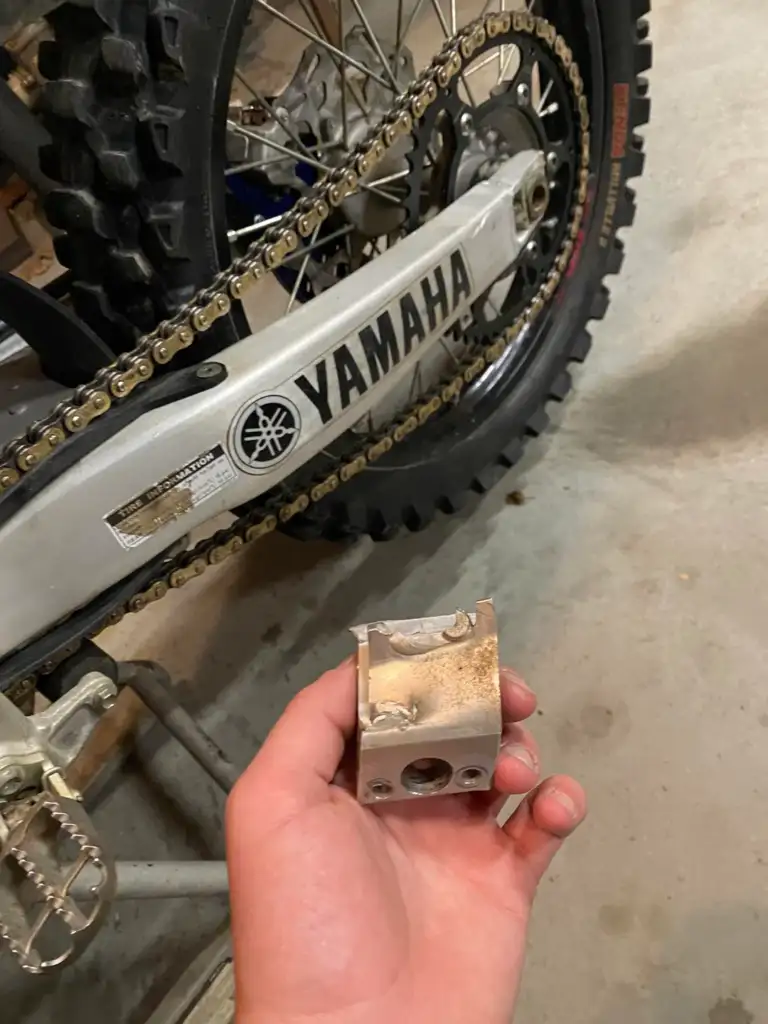 A hand holding a small, rectangular metal part in front of a Yamaha dirt bike's rear wheel. The bike's chain and tire are visible, along with the garage floor.