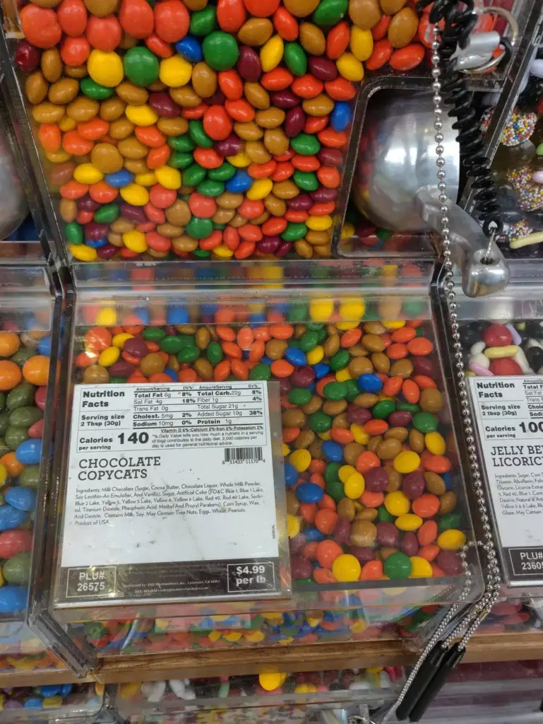 Dispensers filled with colorful candy-coated chocolates labeled "Chocolate Copycats." Nutrition facts indicate 140 calories per serving. Price is $4.99 per pound. A metal scoop on a chain is attached to the display.