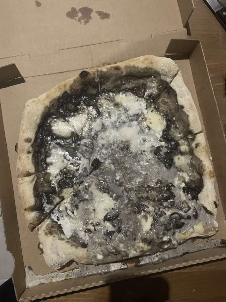 A pizza with a charred crust and toppings that include blackened mushrooms and melted cheese, inside an open cardboard box on a wooden surface.