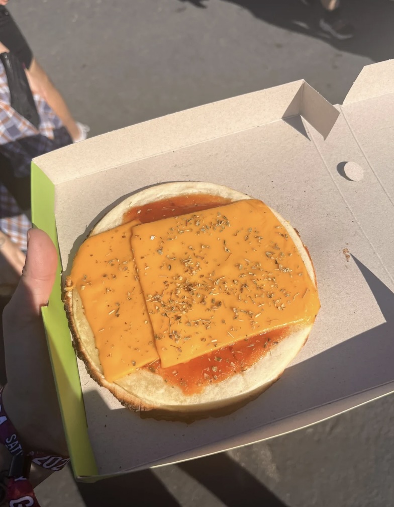 A person holds an open cardboard container with a piece of bread topped with two slices of cheese, some sauce, and sprinkled herbs. Part of a person's arm with a wristband is visible in the sunlight.