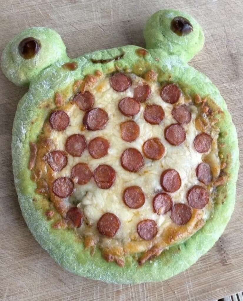 A pizza with a playful design resembling a frog, featuring a green crust and topped with pepperoni slices and melted cheese. The pizza has two round protrusions at the top mimicking eyes.