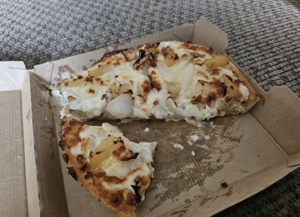 A partially eaten pizza sits in a cardboard box on a textured surface. The pizza is topped with melted cheese, chunks of pineapple, and slices of onion. A slice has been removed, revealing the crust and toppings.