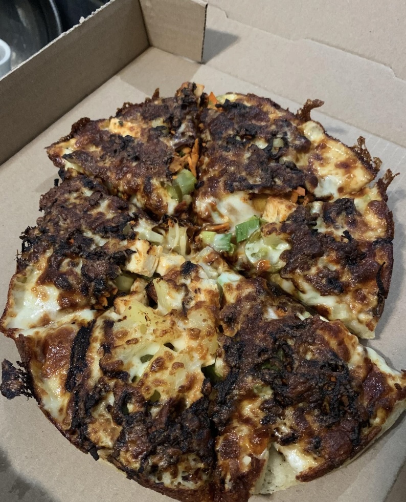 A pizza with a crispy, charred top sits in a cardboard box. It has visible toppings, including melted cheese and green pepper pieces, and is cut into slices. The crust and cheese appear well-done.