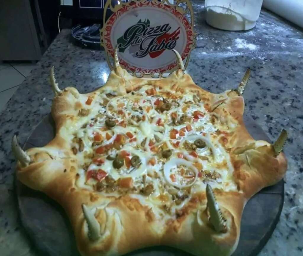 A uniquely shaped pizza with a thick, wavy crust resembling tentacles, topped with cheese, olives, tomatoes, and sausage. A decorative sign on top reads "Pizza do Gabo." The pizza is placed on a marble countertop.