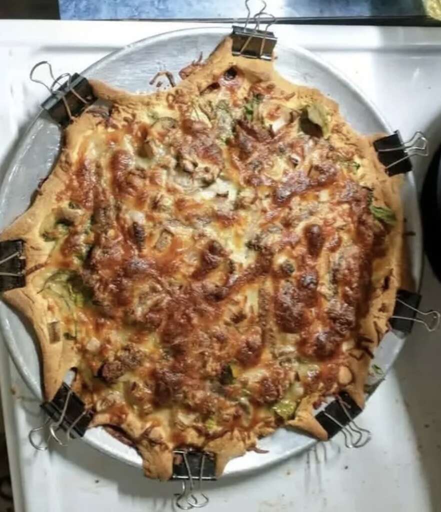 A homemade pizza with a golden crust is held together by binder clips around the edges. The pizza is topped with melted cheese, mushrooms, and green vegetables on a round metal tray.