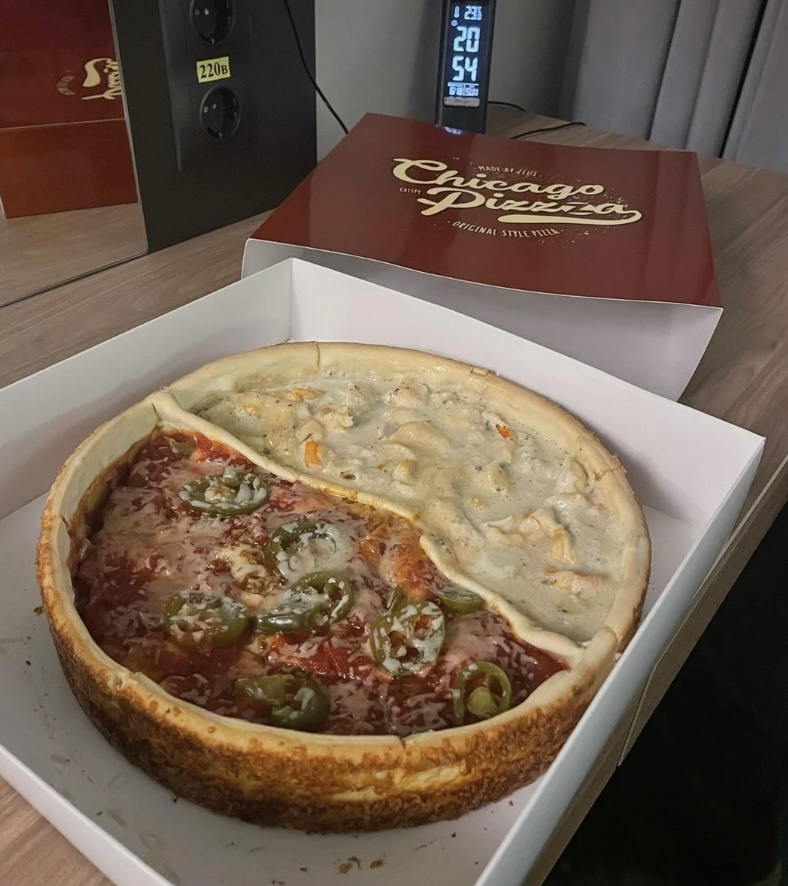 A Chicago-style deep-dish pizza sits open in a box on a wooden table. The pizza is half topped with jalapeños, cheese, and tomato sauce, and half with a creamy sauce and cheese. A digital clock in the background displays 20:54.