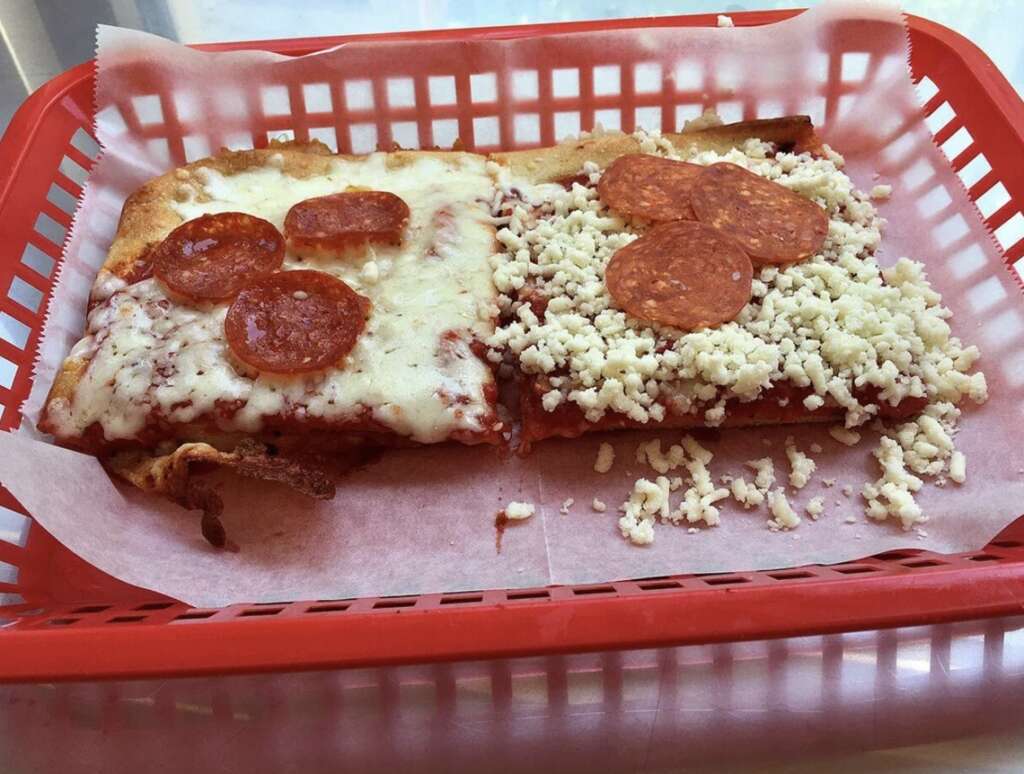 Two square slices of pizza in a red basket lined with parchment paper. One slice has melted cheese and pepperoni, the other has uncooked cheese and pepperoni. Both have crispy edges.