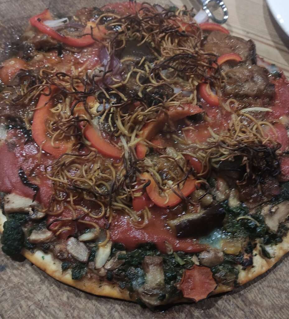 A close-up of a colorful vegetable pizza topped with red peppers, mushrooms, and crispy noodle-like toppings. It has a thin crust and is served on a wooden board.